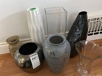 Seven decorative vases are shown arranged on a wooden floor against a white wall, showcasing different materials, sizes, and colors.