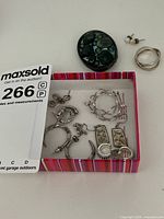 Top down photo of five pairs of sterling silver earrings and one brooch, displayed in a shallow pink and white box with auction lot number card.
