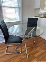Two black leather-like chairs and round glass top table with metal base positioned in bright room with wooden flooring.