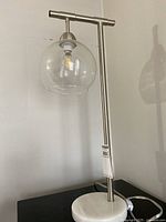 Full view of table lamp with metal vertical stand, T-shaped horizontal top, round white stone-like base, and clear glass globe shade.