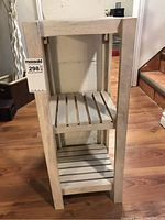 Front view of white wooden three-tier shelf showing slatted shelves and solid vertical supports against a stone wall background on wood floor.