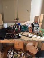 Photo of a wooden workbench with various tools including hammers, pliers, a Bosch drill, boxes of screws and nails, hardware pieces, and a white garbage bin underneath.