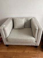 Front view of the off white upholstered arm chair showing the cushioned seat, one loose side cushion, and solid wood legs on a wooden floor.