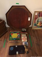 Photo showing vintage octagonal wooden crokinole board standing on the floor next to a chest, with various classic games arranged at its base including Chinese checkers, Hi-Q puzzle, cribbage board, playing cards, pick up sticks, dominoes, wooden checkers and Scrabble set.
