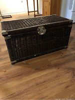 Closed view of the brown wicker chest showing the woven texture, metal corners and central latch on a wooden floor.