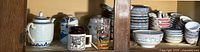 Cabinet shelf showing white and blue ceramic tea set, decorative mugs, glass tumbler, and stacks of various small bowls.