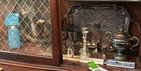 Shelf display of various tarnished silver-plated items including trays, candlesticks, goblets, a decorative urn, and cleaning products in boxes.