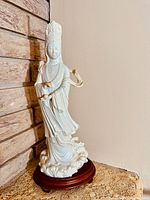 Full side view of tall white porcelain figurine of Kuan Yin on wooden base against wall.