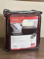 Front view of new Sunbeam Northern Plush heated blanket in original packaging, burgundy color shown.