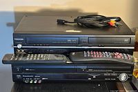 Front view of Toshiba DVD/VCR combo player stacked on Panasonic DMR-EZ49V DVD VCR recorder with three remote controls on top and AV cables coiled on Toshiba unit.