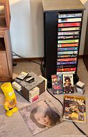 Photo showing the black rotating media console filled with multiple VHS tapes on its shelves, the 2001 8-track cartridge storage box with tapes stacked on top, the sealed vintage Bruce Lee magazine, and the yellow Bart Simpson coin bank placed on the floor next to a vinyl LP album.