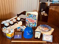 Overview of baseball collectibles on wooden table showing baseballs, Dodgers cap, Babe Ruth plate, book on baseball cards, and gold coin set