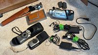 Overview of vintage cameras on carpet including two Samsung digital cameras in cases, a Polaroid-style camera, a small black tripod with brown leather case, Canon digital camcorder, and various cords and power adapters.