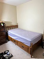 Vintage single wooden bed with mattress on top and three storage drawers underneath positioned against a wall.