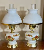 Two vintage milk glass lamps side by side showing floral patterns, brass bases, milk glass shades, and clear glass chimneys