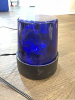 Blue transparent dome rotating flashing light with black base shown on floor surface, close-up showing scratches and wear marking overall condition.