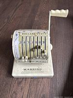 Front angled view of the Paymaster Ribbon Writer showing multiple vertical sliding selectors labeled Dollars and Cents with dollar sign and cent tabs, and a hand crank handle on the right side.