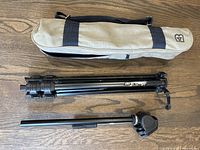 Collapsed Daveco black tripod with beige carrying case placed on wooden floor