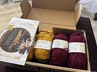 Photo of knitting kit box containing multiple balls of yarn in Hogwarts house colors maroon and gold.