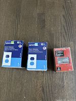 Photo showing two boxes of Leviton plastic wall plates and one red box of Everbilt door hinges with screws on a wooden floor.