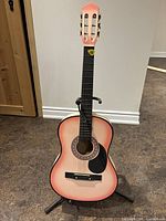 Front view of the pink acoustic guitar standing on a black stand, showing the full body, fretboard, headstock, and attached guitar pick.