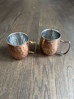Two hammered copper cocktail mugs with rounded bodies and handles shown from above on wooden floor.