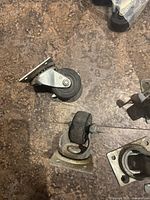 Four swivel casters displayed on a floor surface, showing two with plate mounts and two with stem mounts. The black rubber wheels and silver metal mounts are visible with wear marks.