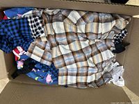 Box containing about 15 men's clothing items with plaid shirts on top in brown and blue tones and other mixed patterns below.
