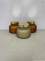 Photo showing set of three new two-wick candles in glass jars; two amber and one beige, all with gold lids.