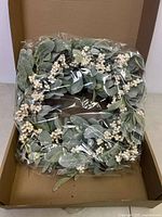 Top view of the square artificial door wreath showing green leaves and white berry clusters, packed in a cardboard box and wrapped in plastic.