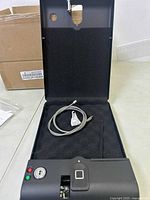 Open fingerprint safe with visible fingerprint sensor, key hole, operation buttons, and attached cable inside with foam padding.