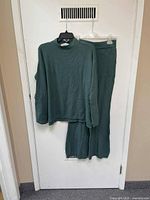 Green knit long-sleeve top and matching jogger pants hanging on hangers against a white door, showing front view of both pieces.