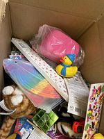 Photo showing inside the box with yellow rubber duck in winter clothing, pink toy purse, colorful craft paper fan, plush cat toy, green cube puzzle, boxed items, and a sheet of emojis stickers.