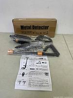 Photo showing metal detector components laid out on a table including main coil sensor, adjustable shaft with orange locking points, armrest, handle, and instruction manual placed in front of original cardboard box labeled 'Metal Detector'.