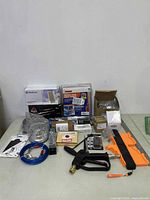 Overview of assorted hardware and tools laid out on table, includes water pipe heat cable box, spray gun handle, blue cable, orange plastic part, and assorted boxes.