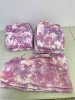 Seven pairs of folded tie-dye jogger pants arranged in three stacks on a plain surface against a white wall.