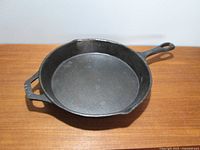 Cast iron frying pan showing top-down view with even wear on cooking surface and two handles, placed on wooden table.