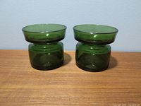 Pair of green glass votive candle holders side by side on a wooden surface, showing front view and shape.