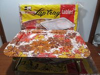 Two vintage fold down lap trays, one opened and one in original packaging, with a floral pattern and metal folding legs.