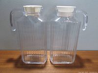 Front view of two rectangular ribbed glass beverage containers with clear handles and white lids on a light background.