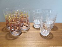 Grouping of four Dominion Glass tinted ombre drinking glasses and five Duralex ornate juice glasses on wooden surface.