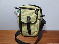 Front view of the light green MEC crossbody bag with black strap, showing front flap pocket with plastic buckle and zippered pocket above.