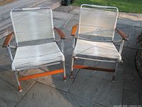 Two vintage folding lawn chairs side by side on stone paving, showing aluminum frames, white plastic tubing seats and backs, wooden armrests and horizontal support bars