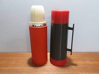 Two insulated beverage containers, one orange Stabilo thermos with ivory lid, one black and red Aladdin Dura-Clad thermos with handle