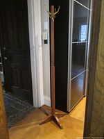Full view of the standing coat rack placed in a corner, showing its height and wooden structure with four splayed feet at the base.