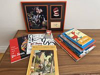 Overview of entire lot on wooden table with framed art at rear and stacks of books and game items in front