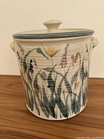 Front view of the stoneware jar showing hand-painted flowers, ventilation hole, two handles, and lid with knob.