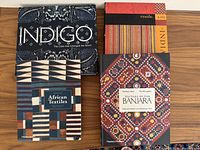 Four textile books arranged flat: 'Indigo: The Colour That Changed the World' (top left), 'Textile Arts of India' (top right), 'African Textiles' (bottom left), 'Textiles of the Banjara' (bottom right).