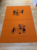 Full view of the orange tablecloth laid flat on a wooden floor showing the repeated black print design of Inuit figures around an igloo.
