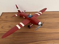Front-side angle of red vintage airplane sculpture with aviator pilot figure sitting in cockpit. Plane has white stripes on wings and tail.